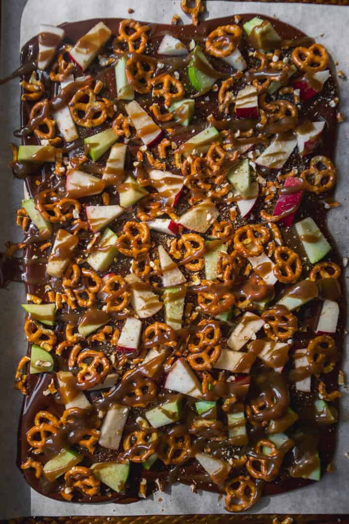 A sheet of chocolate bark topped with chopped apples, mini pretzels, and drizzles of caramel, displayed on parchment paper.