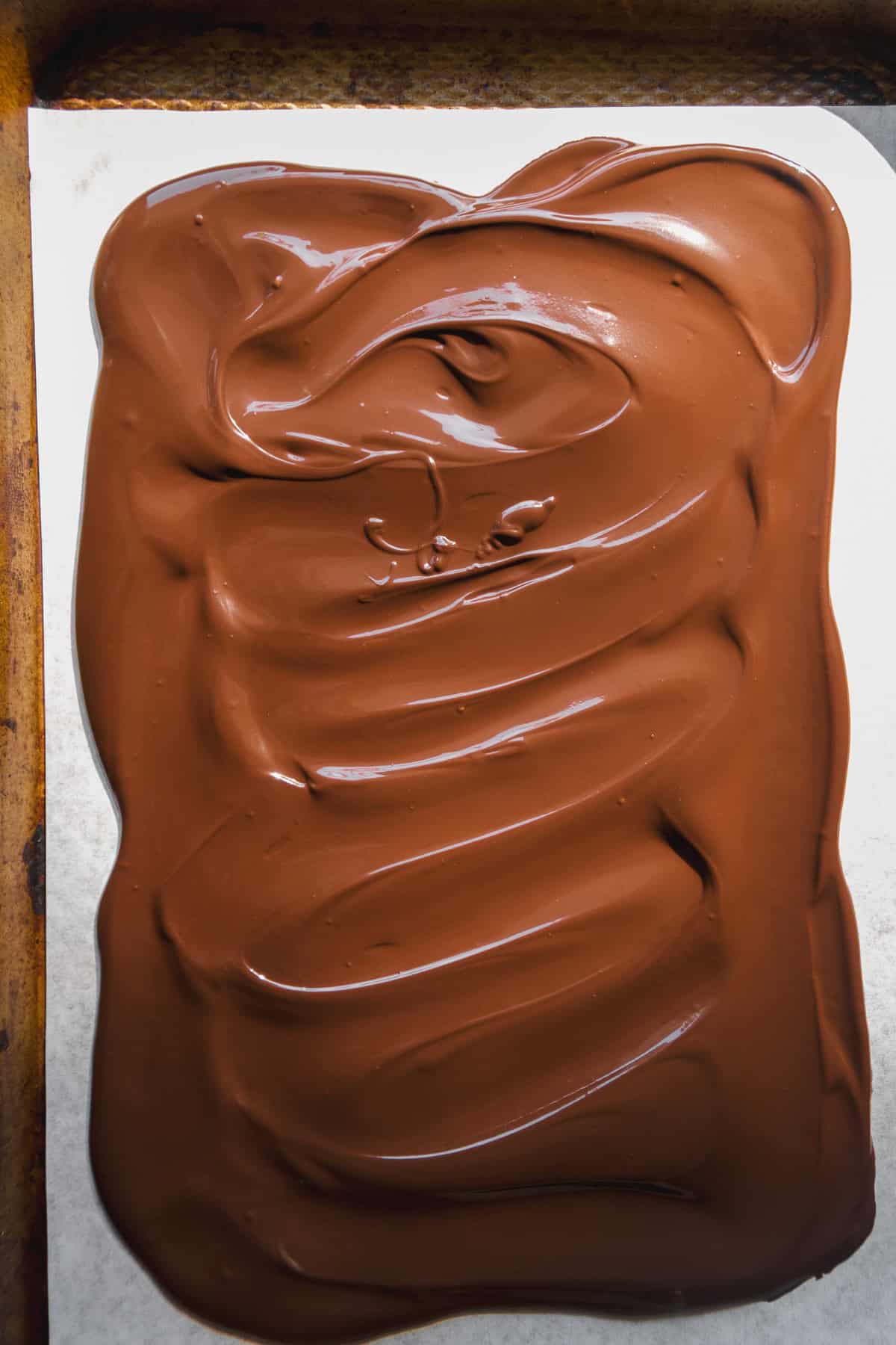 A sheet of smooth, melted milk chocolate is spread evenly on parchment paper, with swirling patterns visible on the surface.