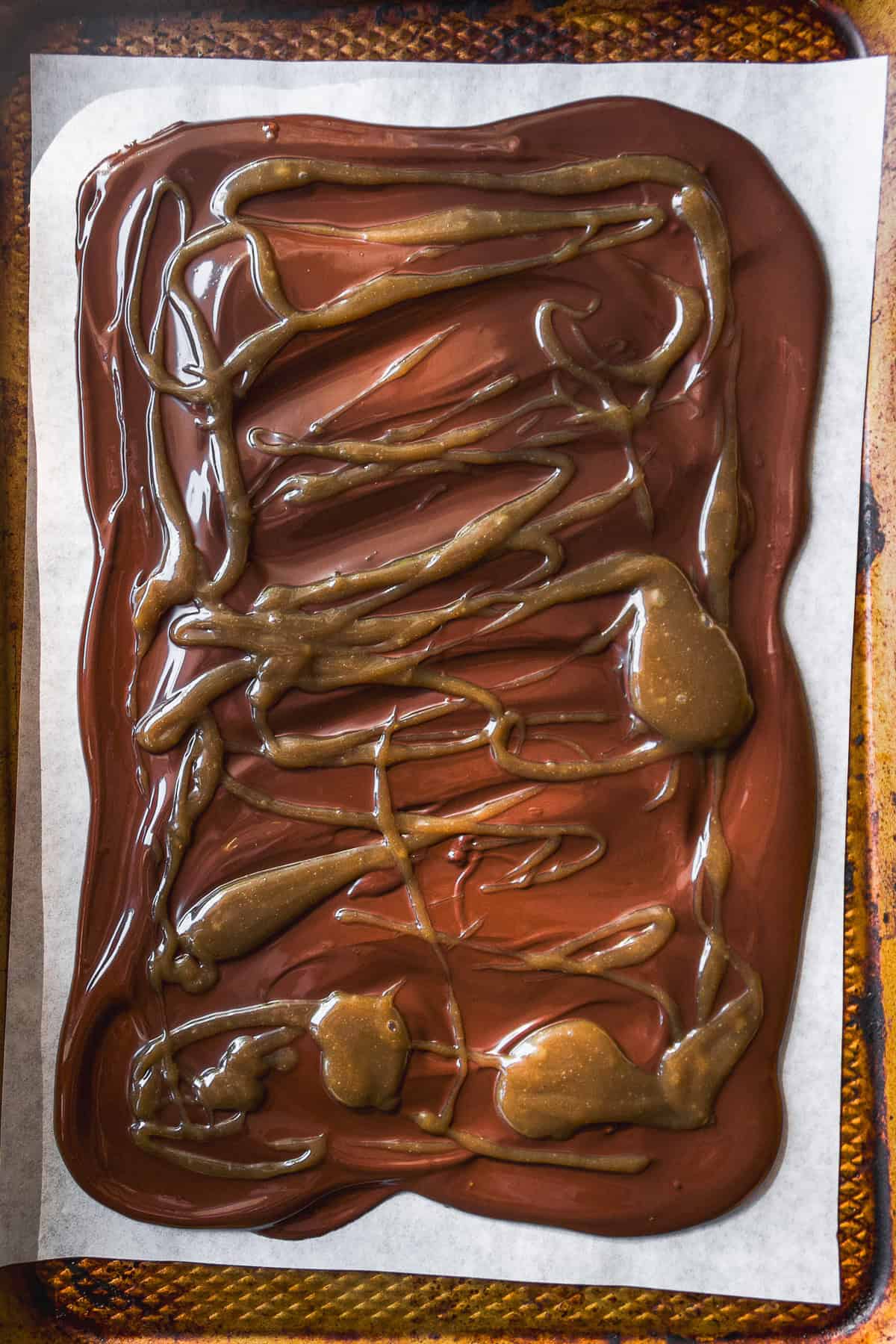A sheet of parchment paper on a baking tray holds a layer of melted chocolate, swirled and topped with drizzles and pools of melted caramel.
