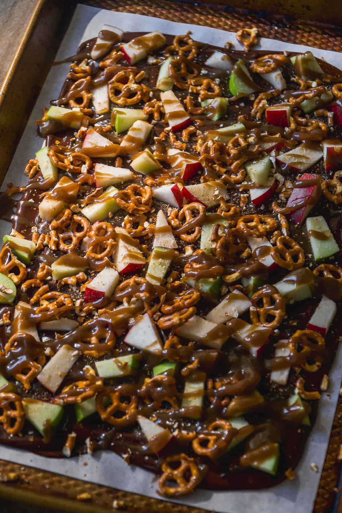 A baking sheet lined with parchment paper holds a layer of chocolate topped with chopped apples, pretzel pieces, and drizzled caramel.