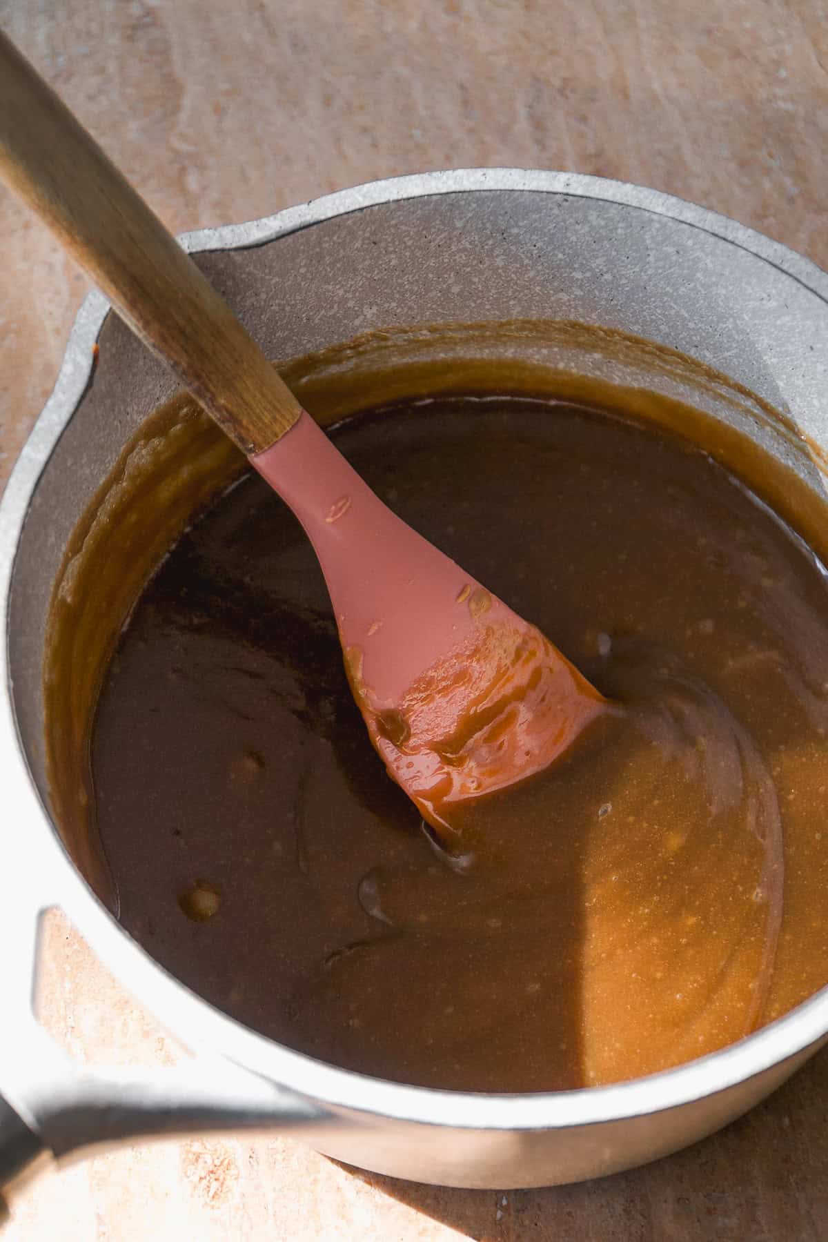 A pink spatula stirs thick, golden caramel sauce in a light gray saucepan sitting on a brown countertop, with sunlight casting a shadow across part of the mixture.