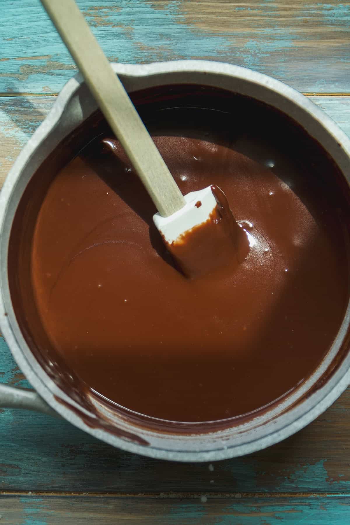 A saucepan filled with smooth, melted chocolate being stirred with a white silicone spatula on a rustic blue wooden surface.