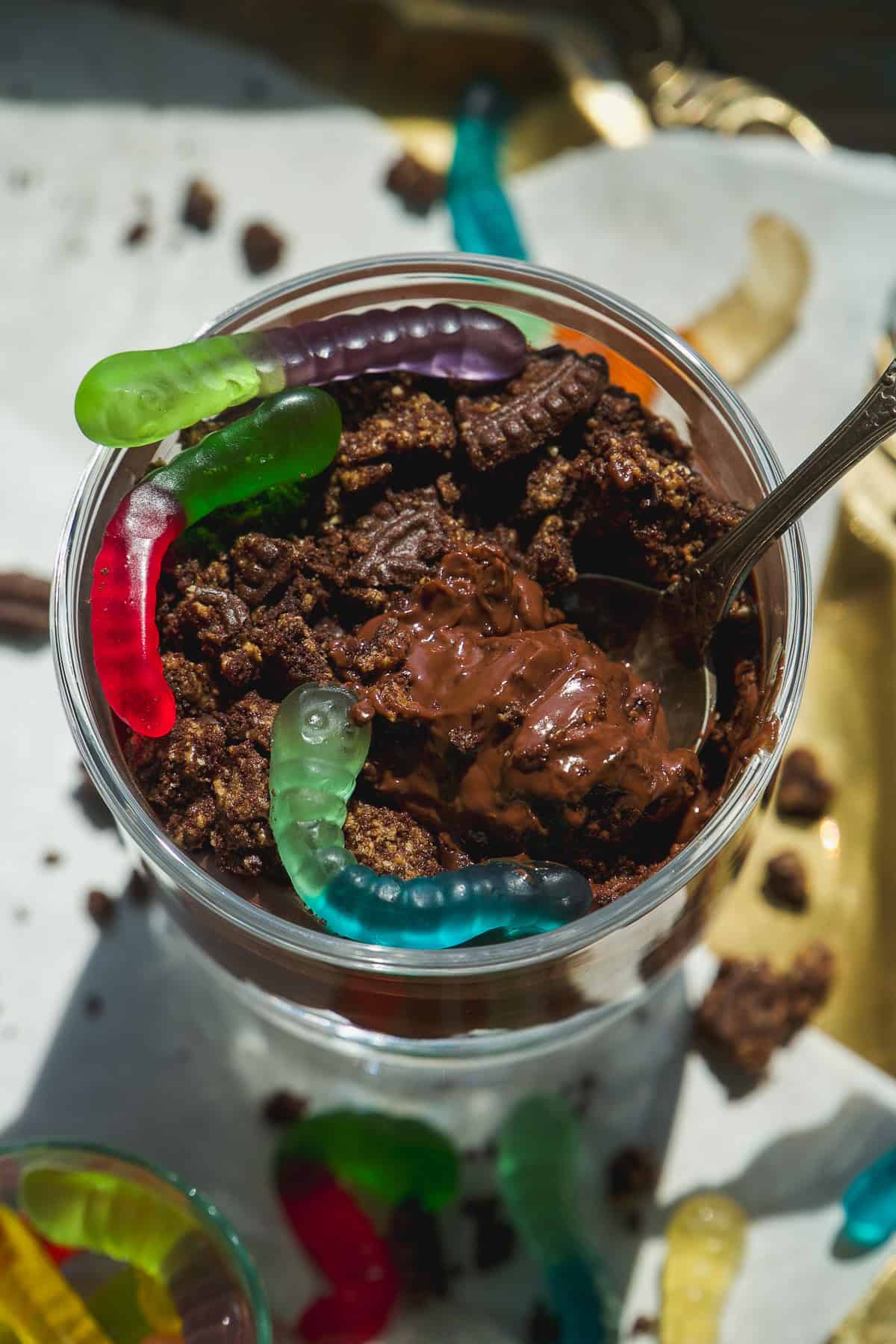 A close-up of a dessert cup filled with chocolate pudding, crumbled cookies, and topped with colorful gummy worms, with a spoon partially inserted into the dessert.