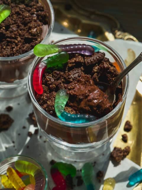 A glass cup filled with chocolate pudding, crumbled cookies, and colorful gummy worms on top, with a spoon sticking out. Another cup and loose gummy worms are visible nearby.