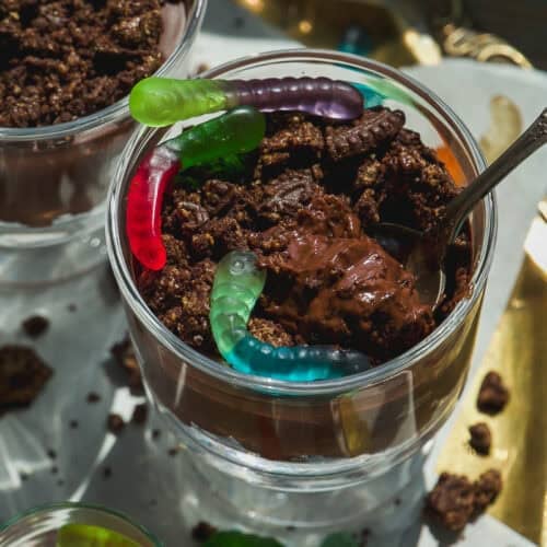 A glass cup filled with chocolate pudding, crumbled cookies, and colorful gummy worms on top, with a spoon sticking out. Another cup and loose gummy worms are visible nearby.