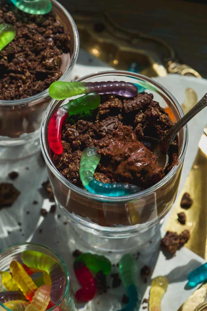 A glass cup filled with chocolate pudding, crumbled cookies, and colorful gummy worms on top, with a spoon sticking out. Another cup and loose gummy worms are visible nearby.