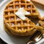 A stack of waffles topped with a pat of melting butter and drizzled with syrup sits on a plate. A fork holds a piece of the waffle, and a knife rests nearby on the wooden table.