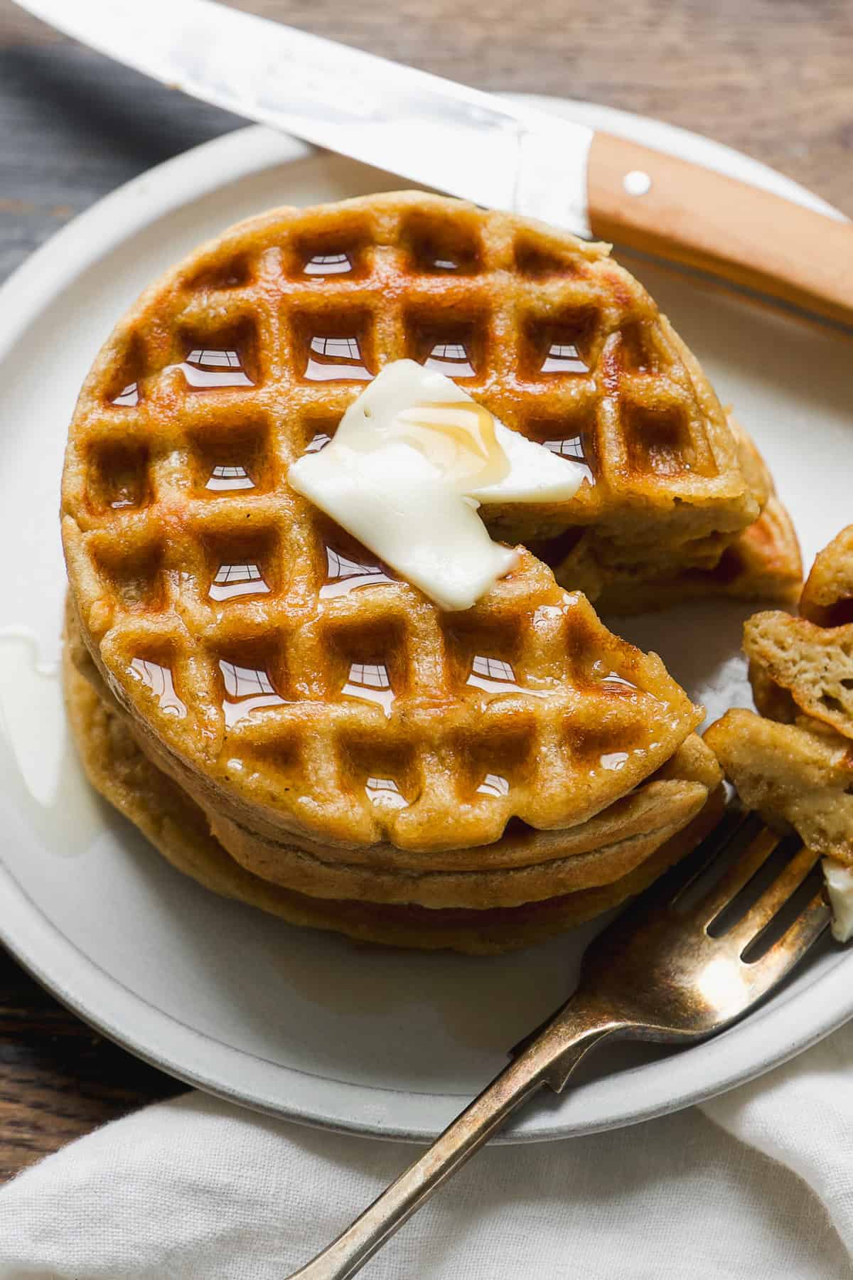 A stack of gluten free waffles topped with a pat of melting butter and drizzled with syrup sits on a plate. A fork holds a piece of the waffle, and a knife rests nearby on the wooden table.