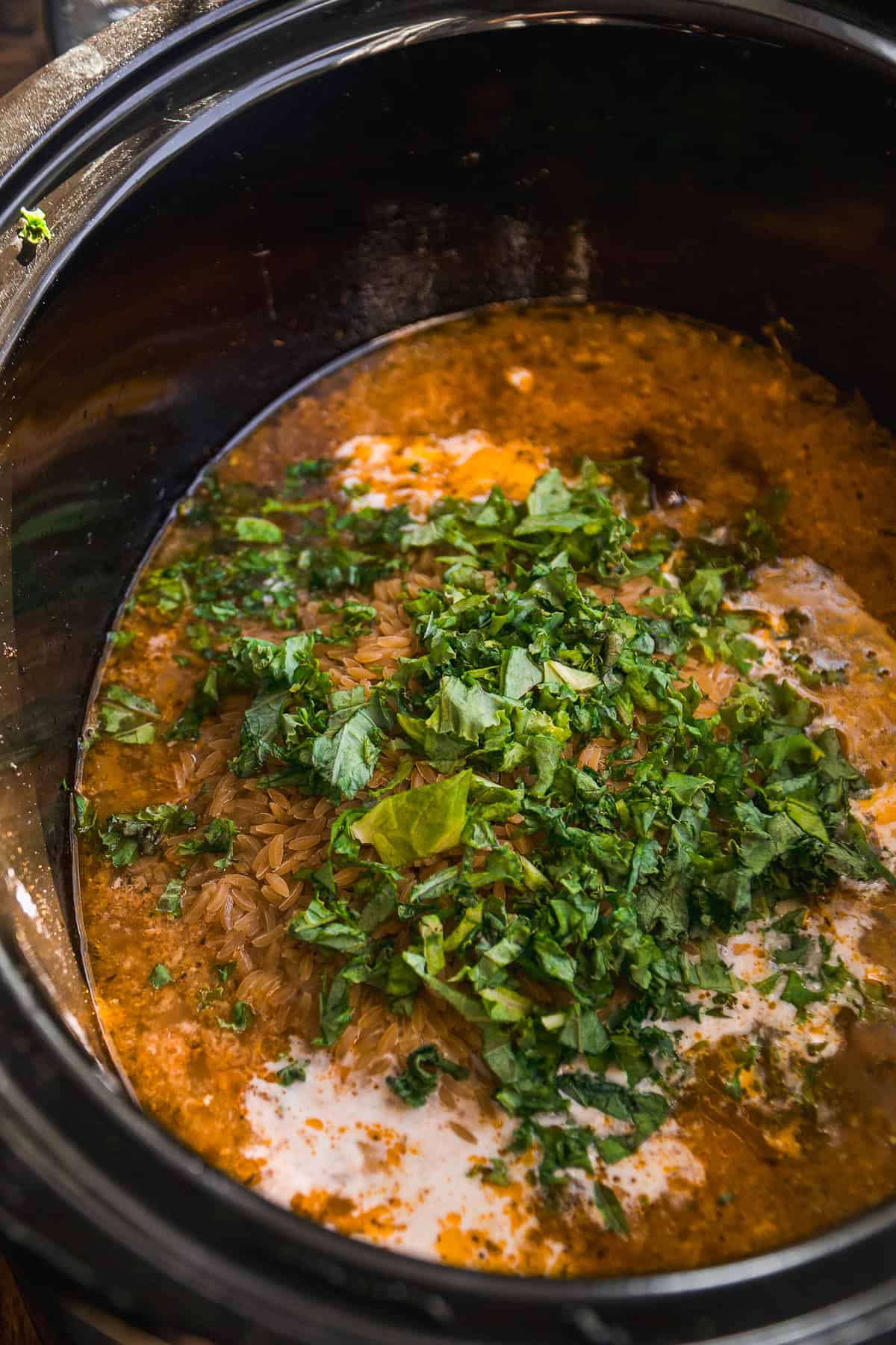 A close-up of a slow cooker filled with a creamy, orange-colored sauce, rice, and chopped fresh cilantro on top, ready to be stirred and cooked.