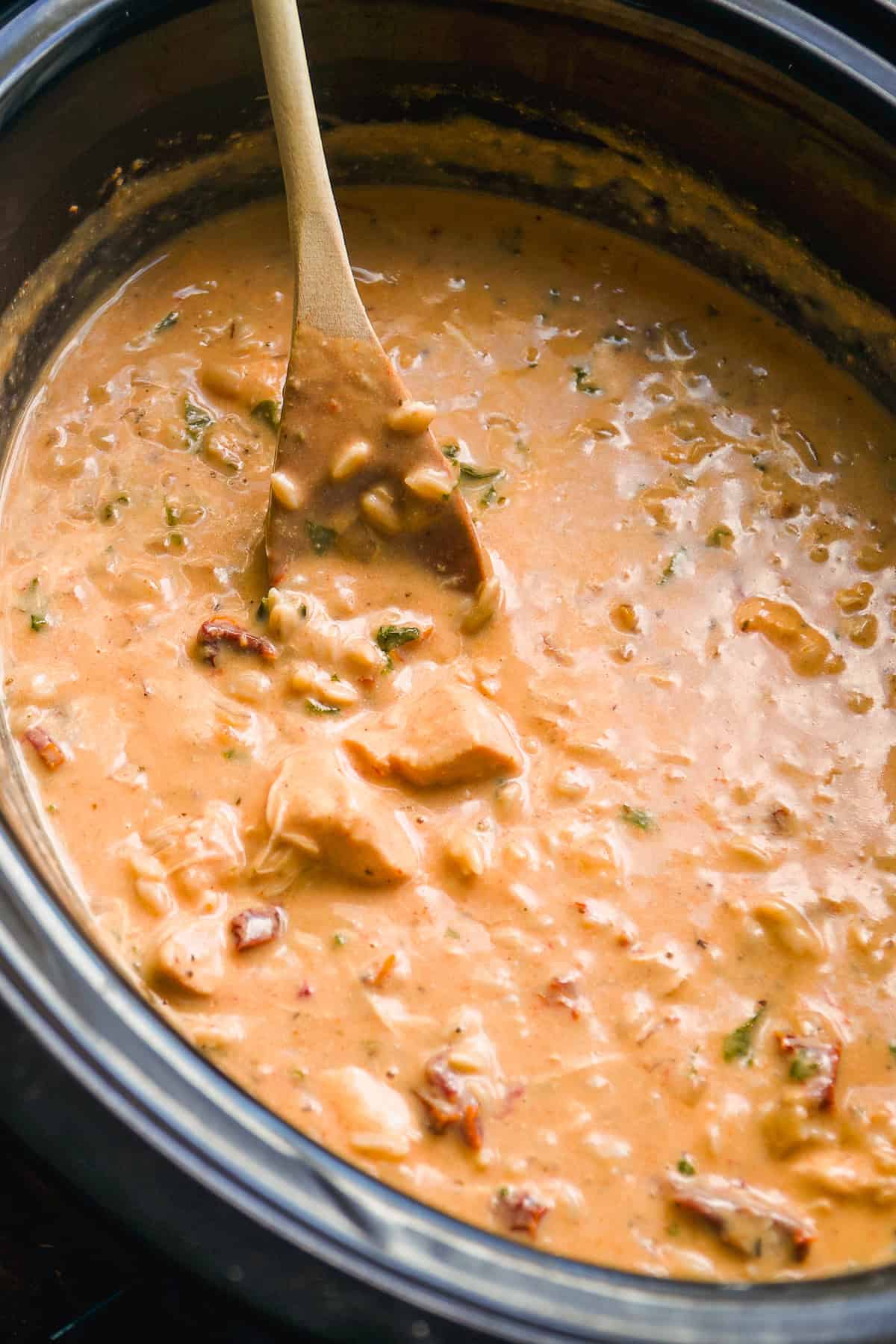 A wooden spoon stirs a thick, creamy soup with chunks of meat and vegetables inside a black slow cooker.