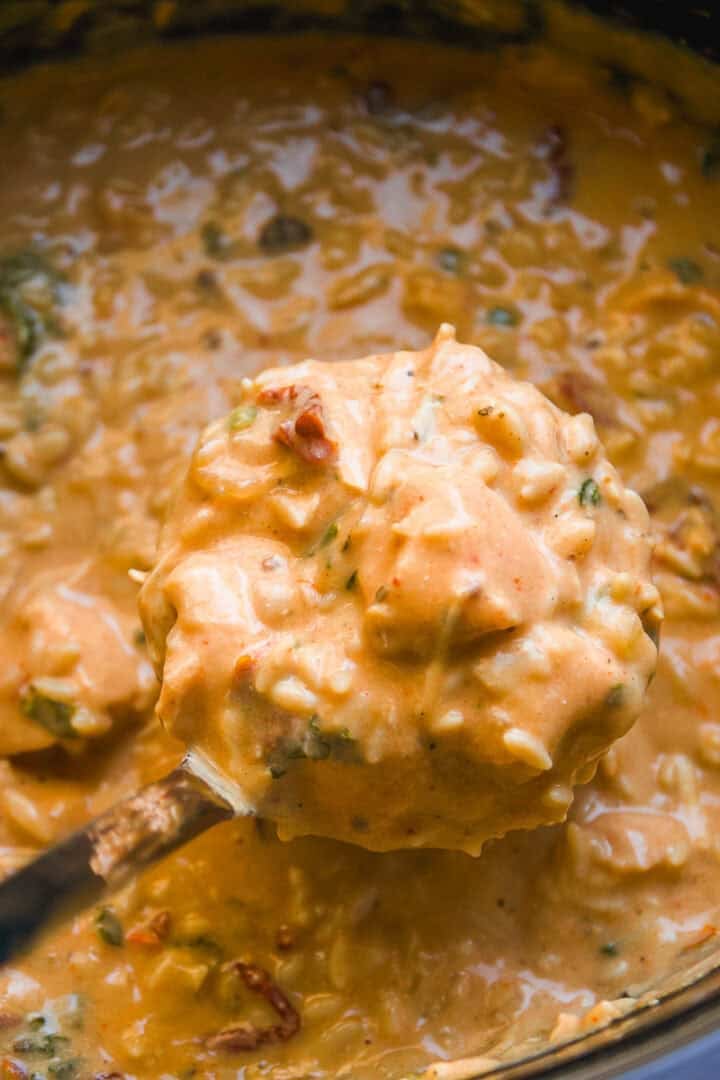 A close-up of a creamy, orange-colored soup or stew with chunks of chicken, rice, herbs, and vegetables being scooped up with a ladle.