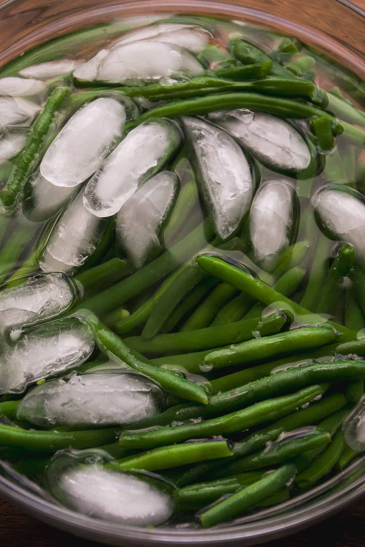 A close-up of fresh green beans submerged in a bowl of ice water with several ice cubes floating on top.
