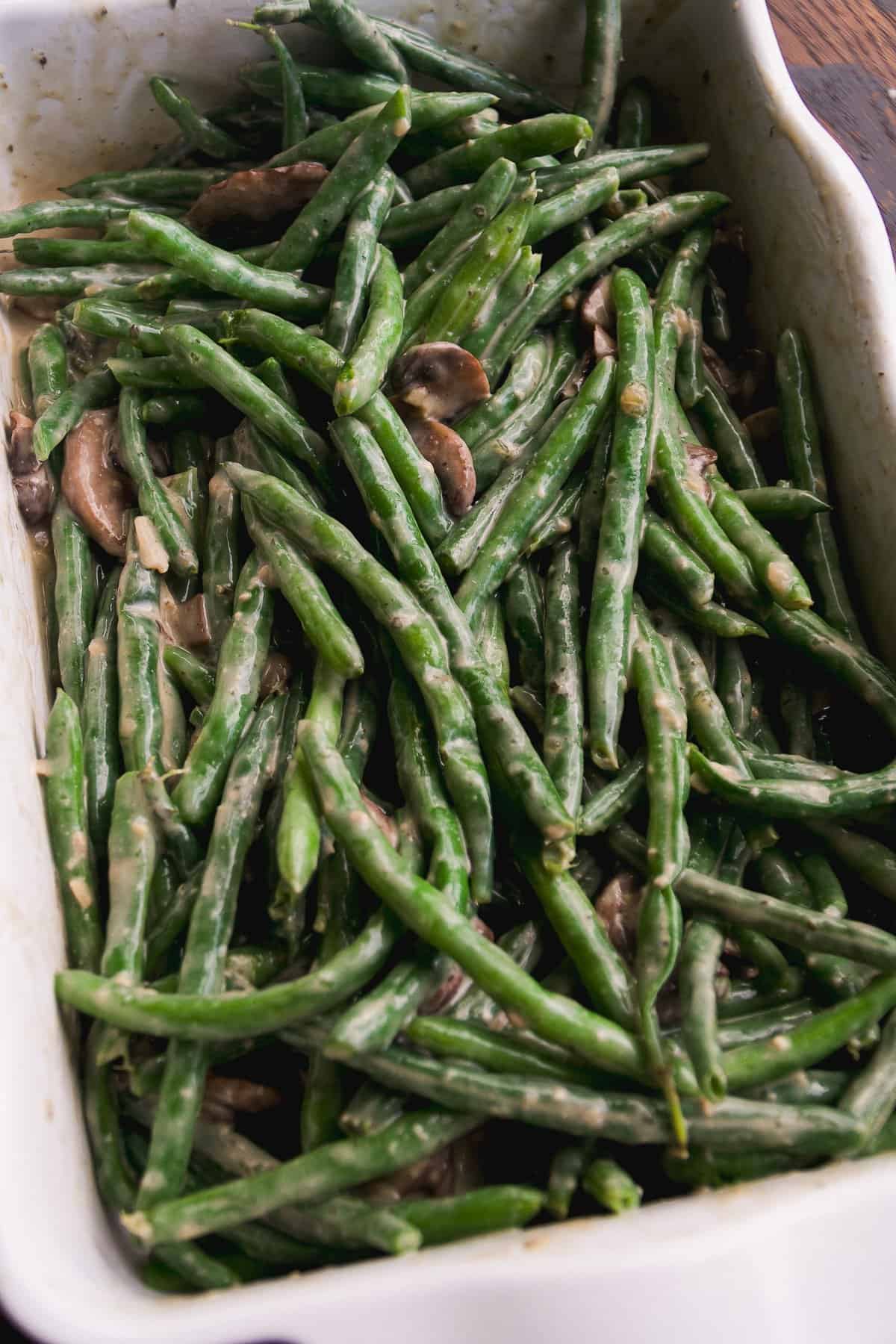 A close-up of a white baking dish filled with green beans and sliced mushrooms coated in a creamy sauce.