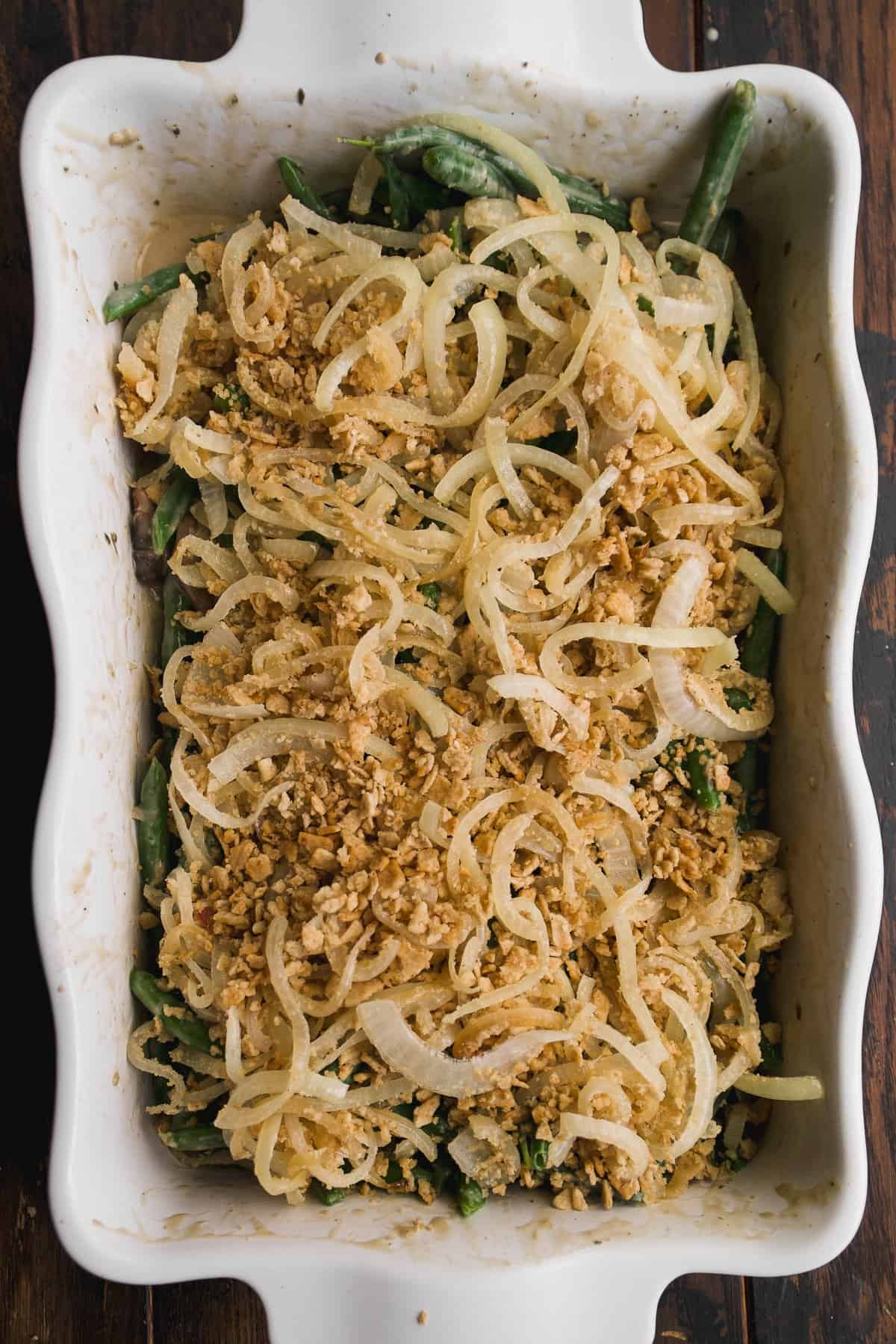 A white baking dish filled with green bean casserole, topped with crispy fried onions and breadcrumbs, sits on a dark wooden surface.