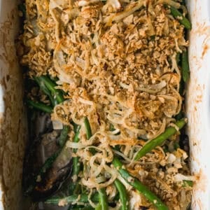 A green bean casserole topped with crispy fried onions in a white baking dish, partially served with a metal spoon resting to the side.