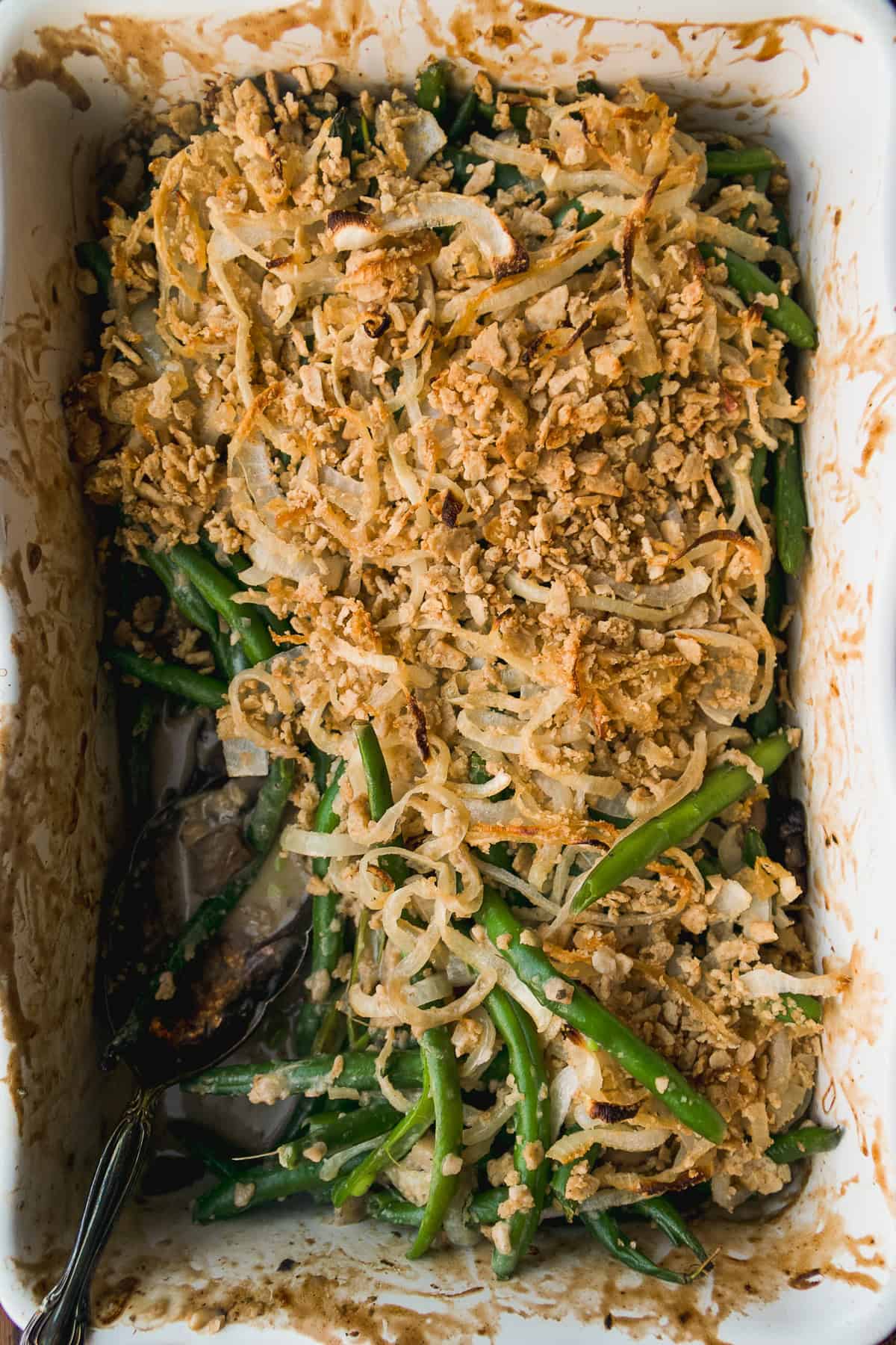 A green bean casserole topped with crispy fried onions in a white baking dish, partially served with a metal spoon resting to the side.