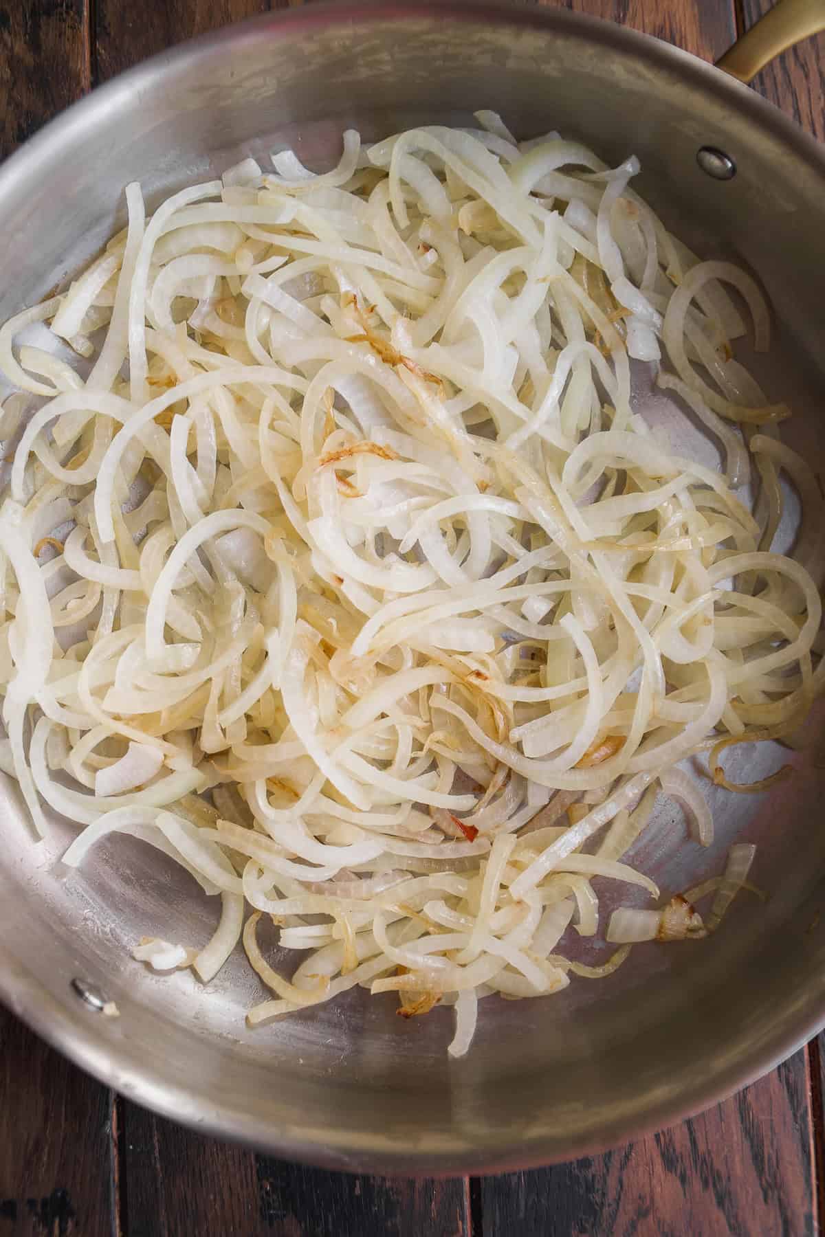 Thinly sliced onions are lightly sautéing in a stainless steel pan, beginning to turn translucent with a few golden edges, on a dark wooden surface.