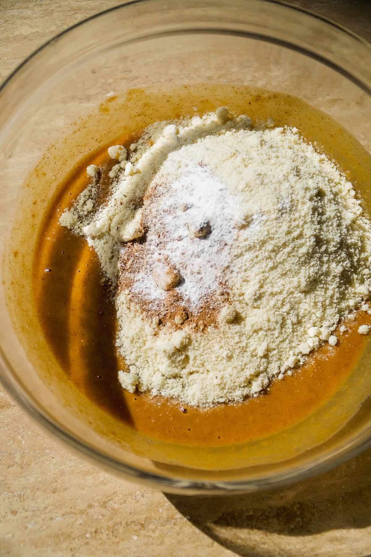 A glass bowl containing a mixture of wet and dry baking ingredients, including a light brown liquid and a mound of flour or almond meal with baking powder, seen from above in natural light.