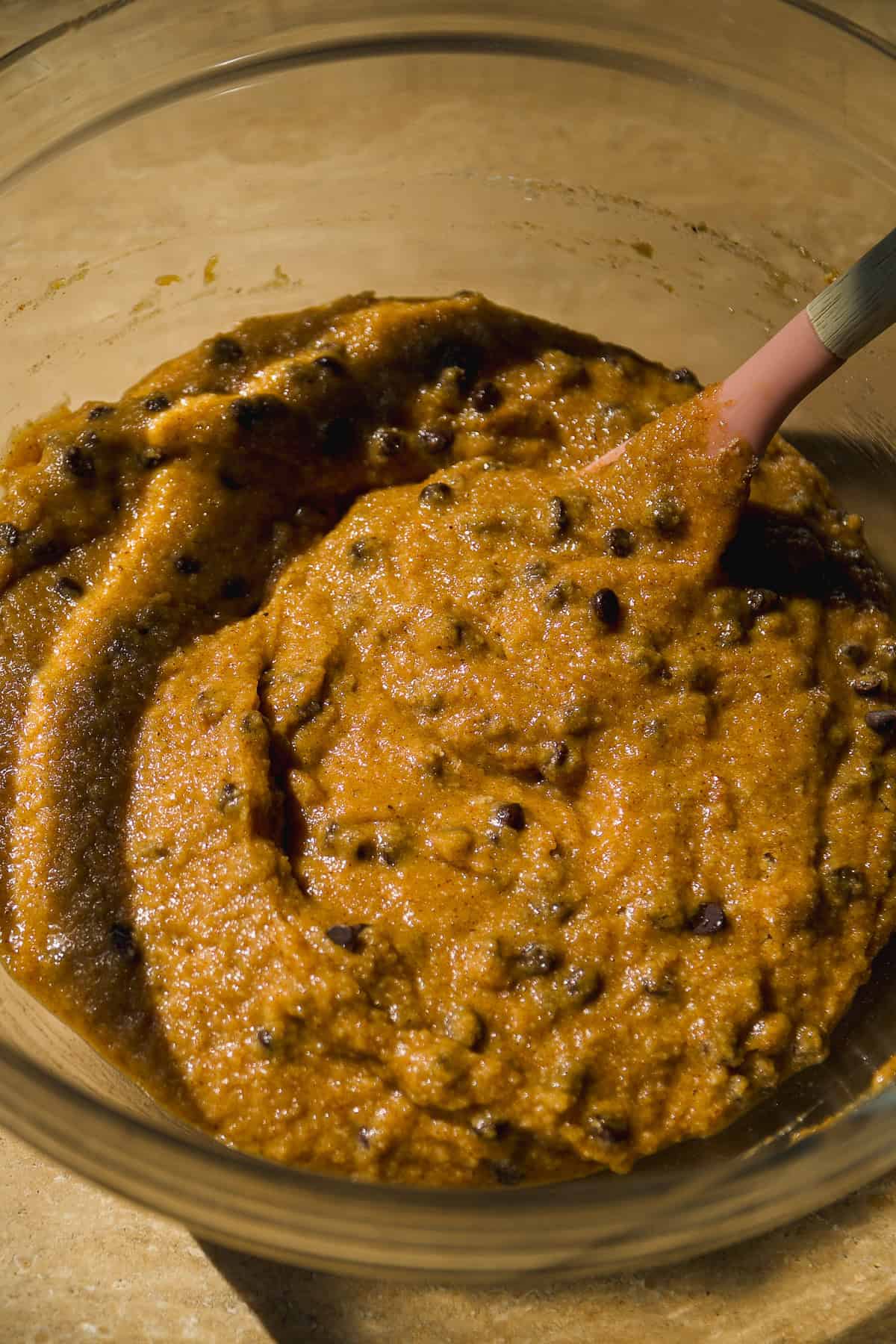 A close-up of a glass bowl containing thick, orange-brown cookie dough with chocolate chips, and a spatula partially submerged in the mixture.