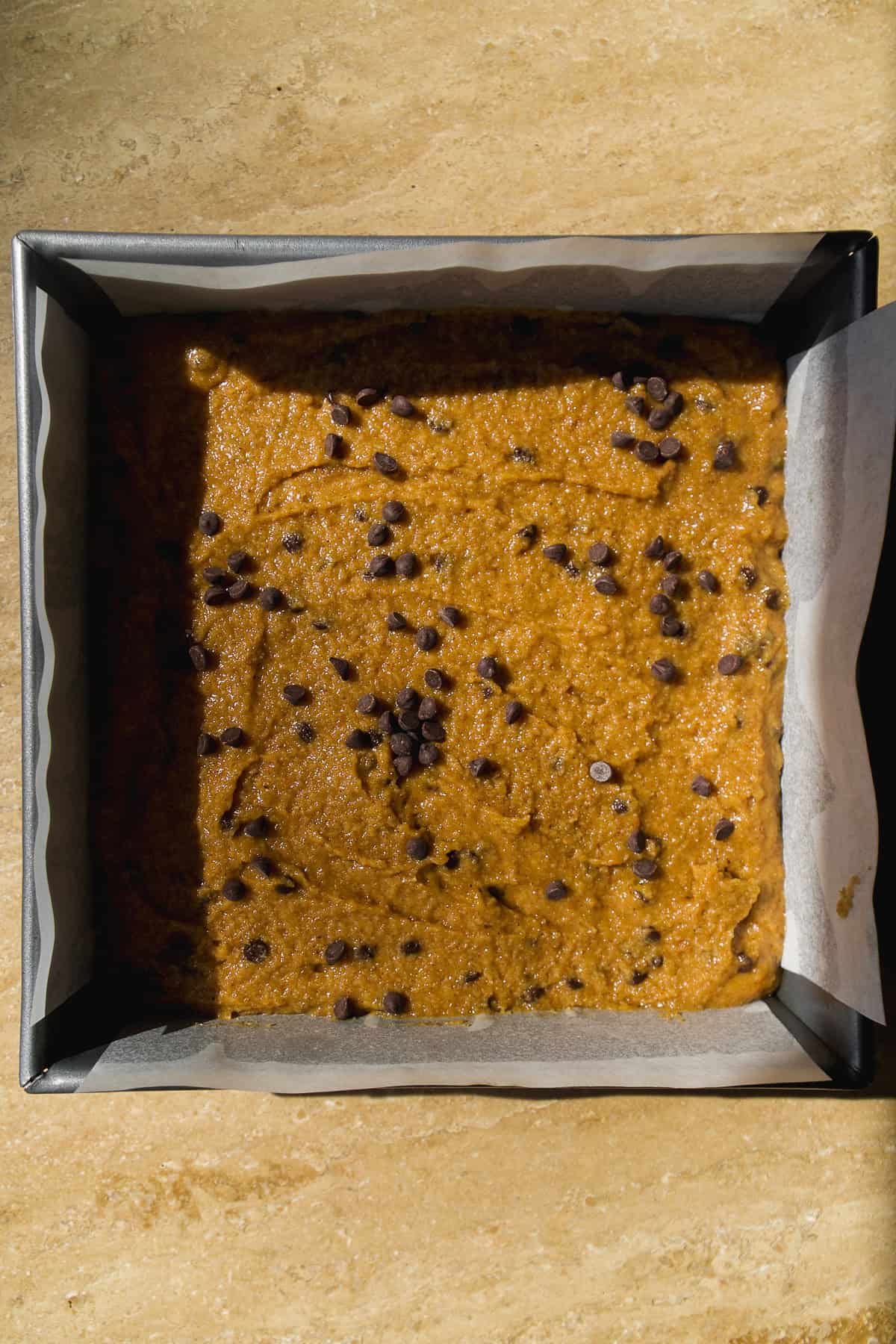 A square baking pan lined with parchment paper is filled with unbaked cookie dough topped with scattered chocolate chips, sitting on a light brown countertop.