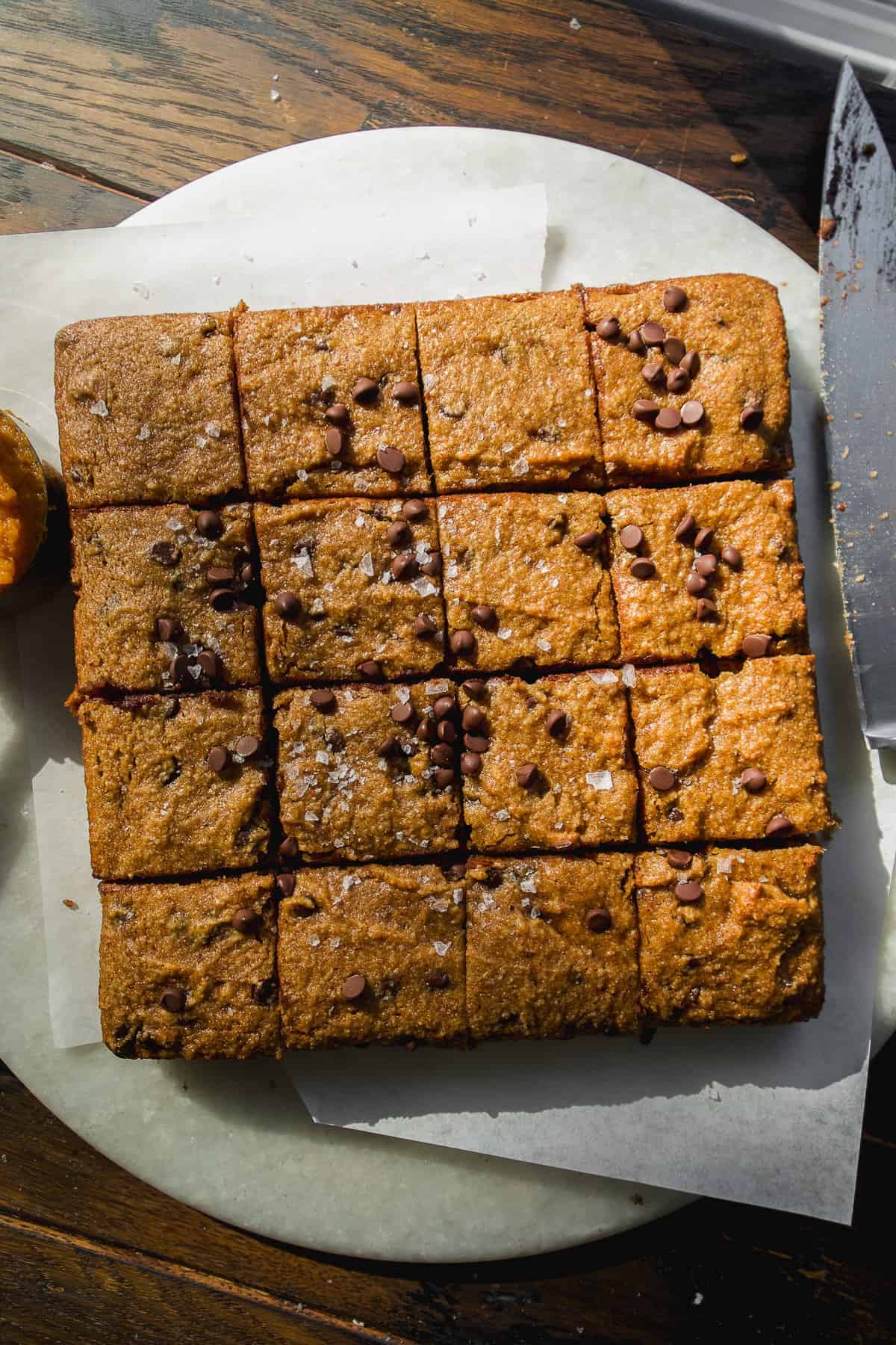 A square pan of baked blondies cut into sixteen pieces, topped with sea salt and scattered chocolate chips, sits on parchment paper on a round marble board with a knife nearby.