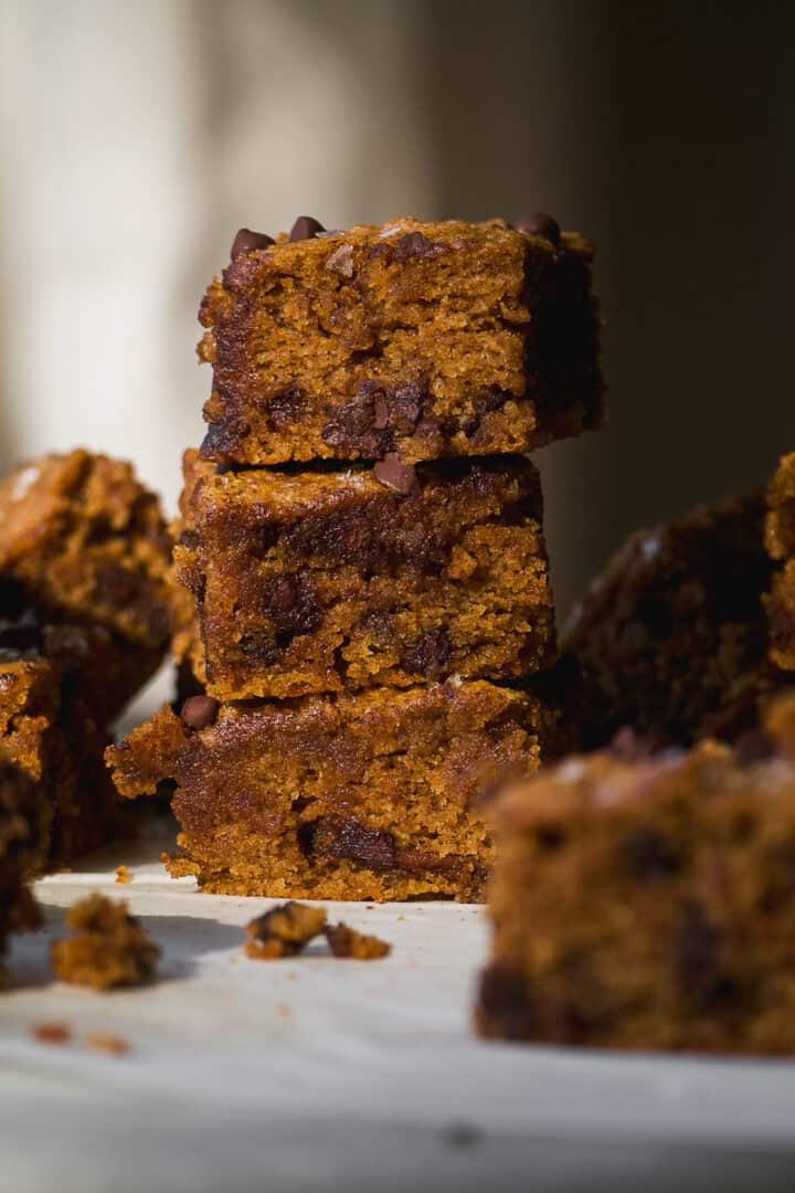 Three stacked chocolate chip blondies with a moist, crumbly texture are in focus, surrounded by more blondie pieces on a light surface. The lighting highlights their golden-brown color and scattered chocolate chips.