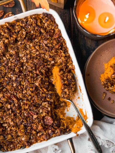 A baked sweet potato casserole topped with a crunchy oat and pecan streusel sits in a white dish. A serving has been scooped out with a spoon. Nearby are a lit candle and a brown plate on a dark wooden table.