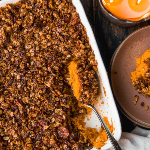 A baked sweet potato casserole topped with a crunchy oat and pecan streusel sits in a white dish. A serving has been scooped out with a spoon. Nearby are a lit candle and a brown plate on a dark wooden table.