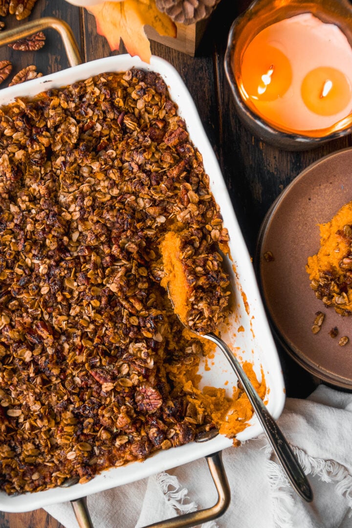 A baked sweet potato casserole topped with a crunchy oat and pecan streusel sits in a white dish. A serving has been scooped out with a spoon. Nearby are a lit candle and a brown plate on a dark wooden table.