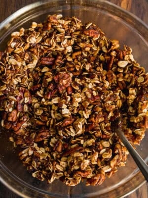 A glass bowl filled with a mixture of oats, chopped pecans, and spices, with a spoon resting inside, on a wooden surface.