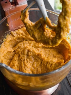 A close-up of orange batter being mixed in a stand mixer bowl, with some batter clinging to the mixer attachment above the bowl. The bowl and mixer sit on a dark wooden surface.