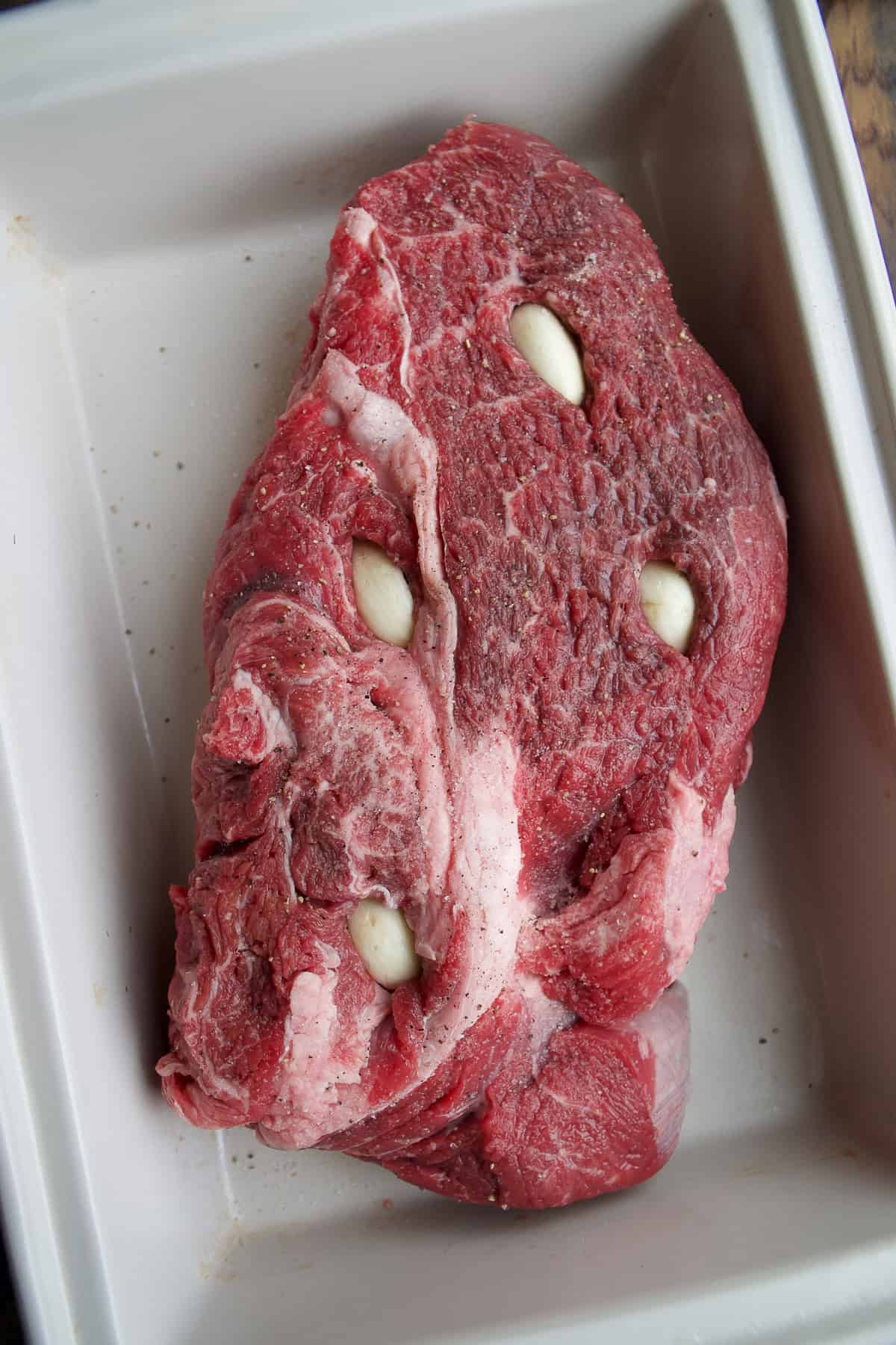 A raw beef roast sits in a white baking dish with several garlic cloves inserted into slits in the meat.