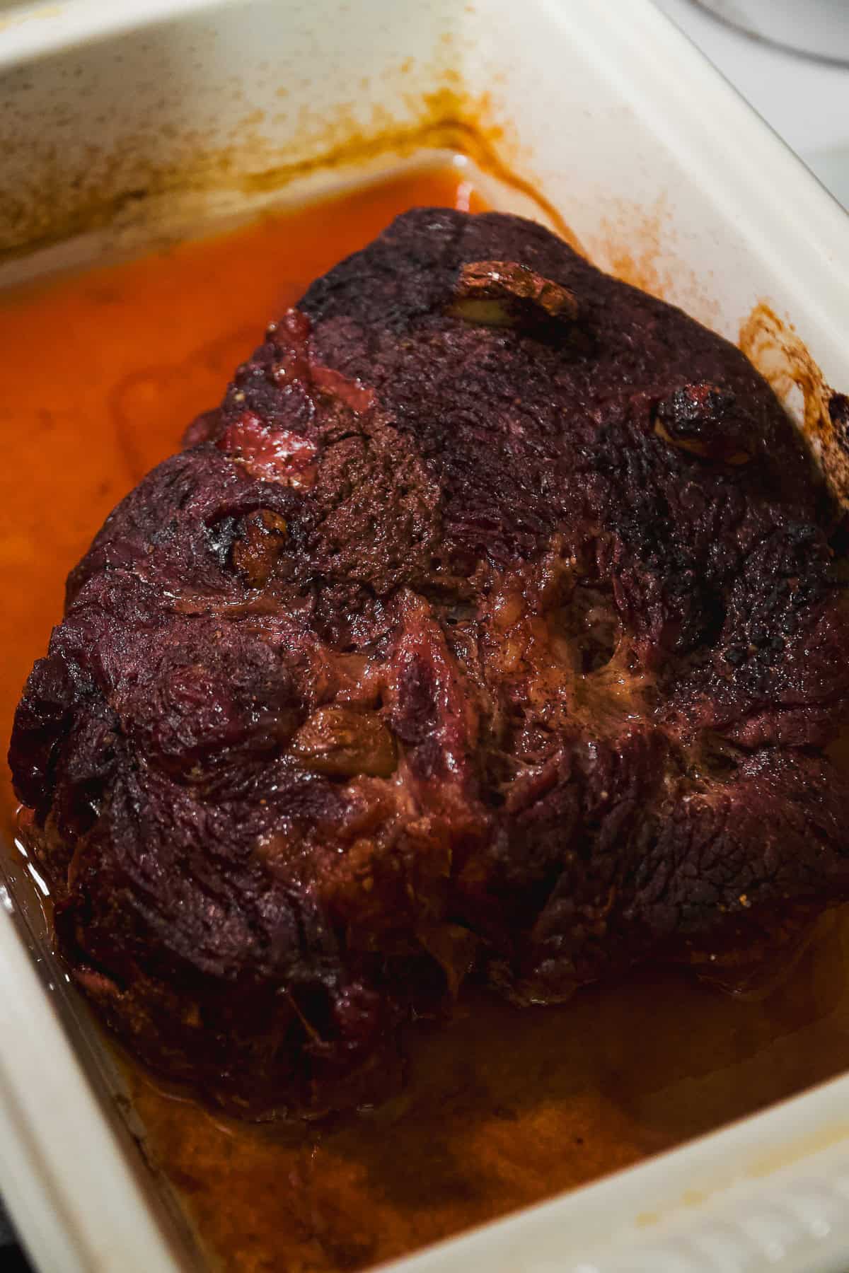 A large, cooked beef roast with a dark, browned crust sits in a white baking dish, surrounded by a pool of reddish-brown juices.