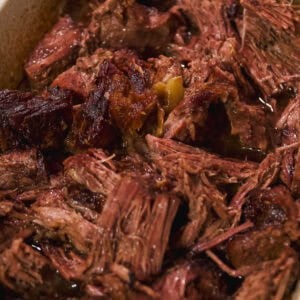 Shredded, juicy roast beef sits in a white baking dish, with visible tender pieces and rich brown juices pooling at the bottom.