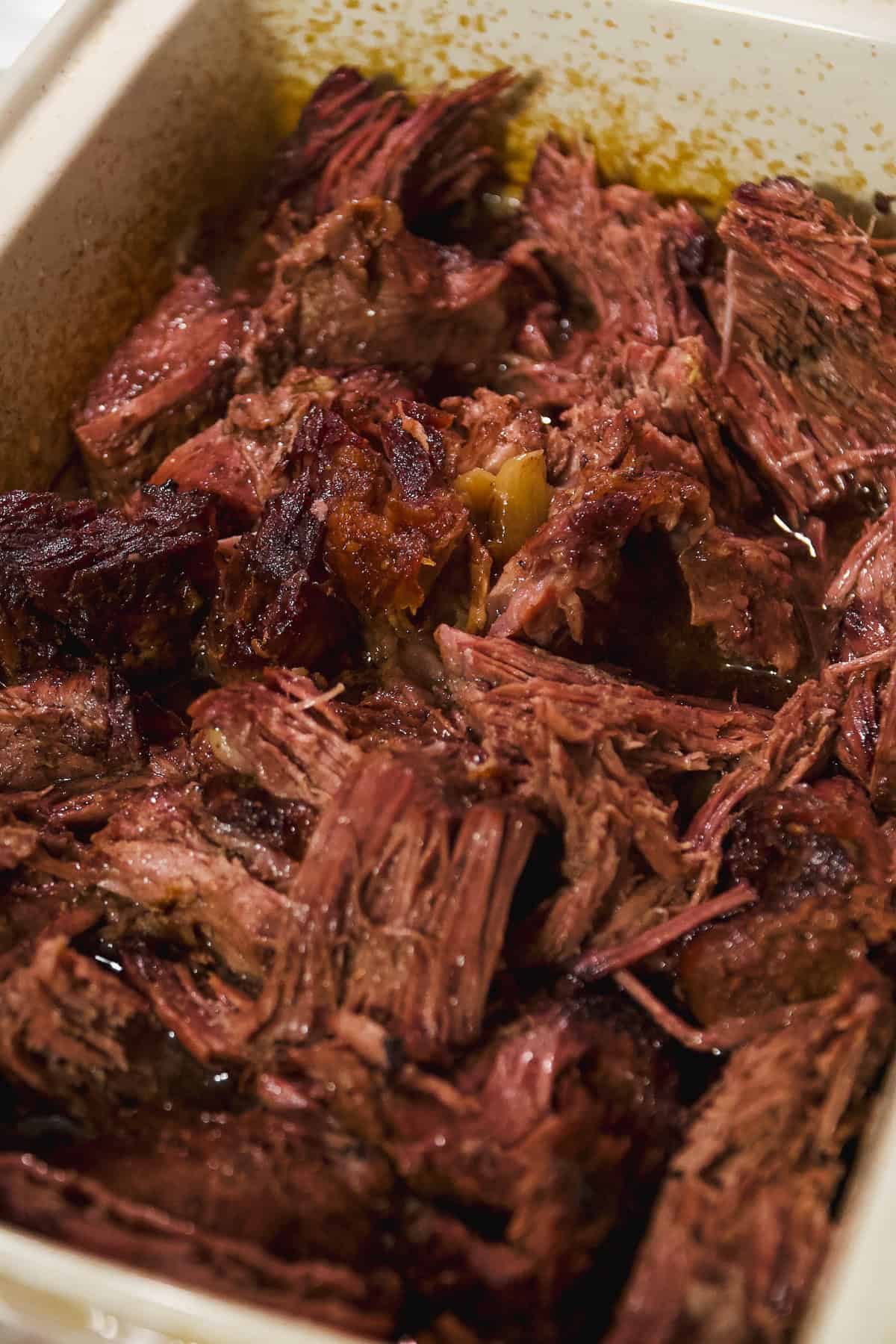 Close-up of tender, shredded pot roast beef in a white baking dish, with visible juices and browned edges.