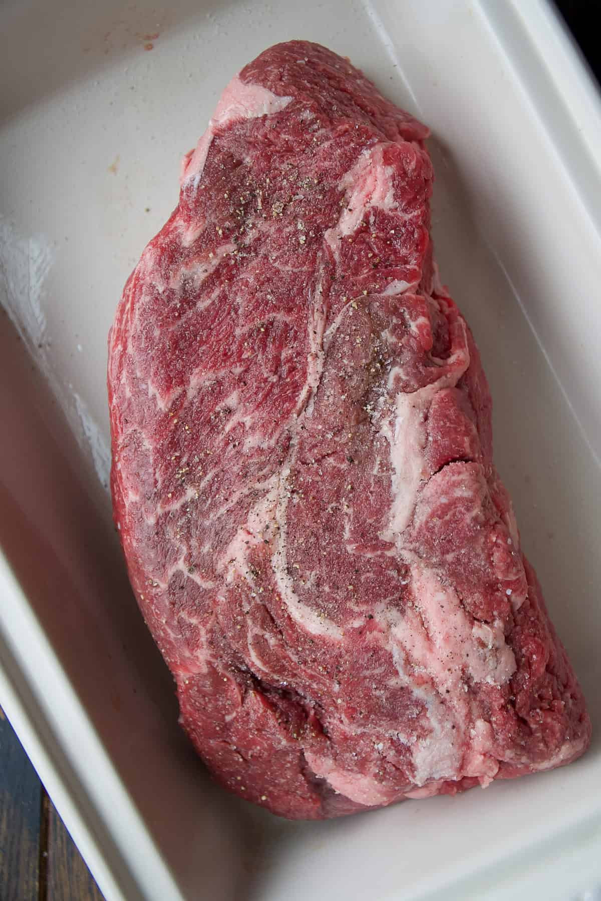 A raw, marbled beef roast with visible fat is seasoned with salt and pepper and placed in a white rectangular baking dish.
