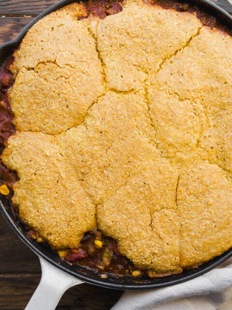 A cast iron skillet filled with chili, topped with a golden, crumbly cornbread crust, sits on a wooden surface with a white cloth partially visible underneath.