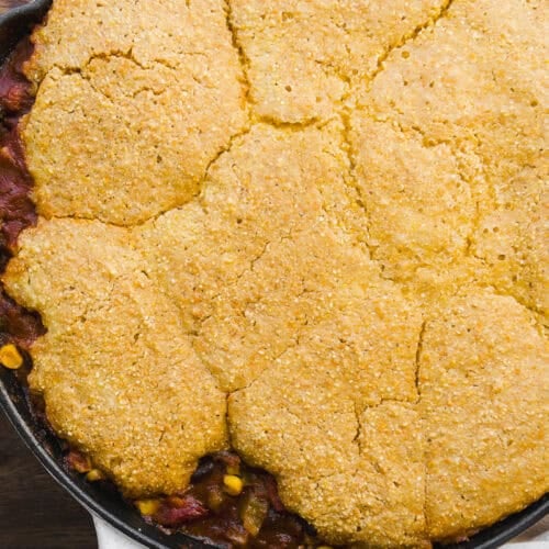 A cast iron skillet filled with chili, topped with a golden, crumbly cornbread crust, sits on a wooden surface with a white cloth partially visible underneath.