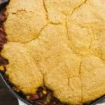 A skillet filled with chili topped with a golden, cracked cornbread crust, sitting on a wooden surface with part of a white cloth visible under the handle.