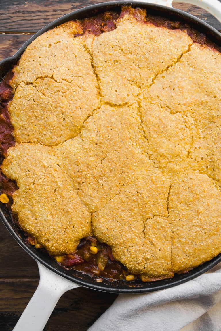 A cast iron skillet filled with chili, topped with a golden, crumbly cornbread crust, sits on a wooden surface with a white cloth partially visible underneath.