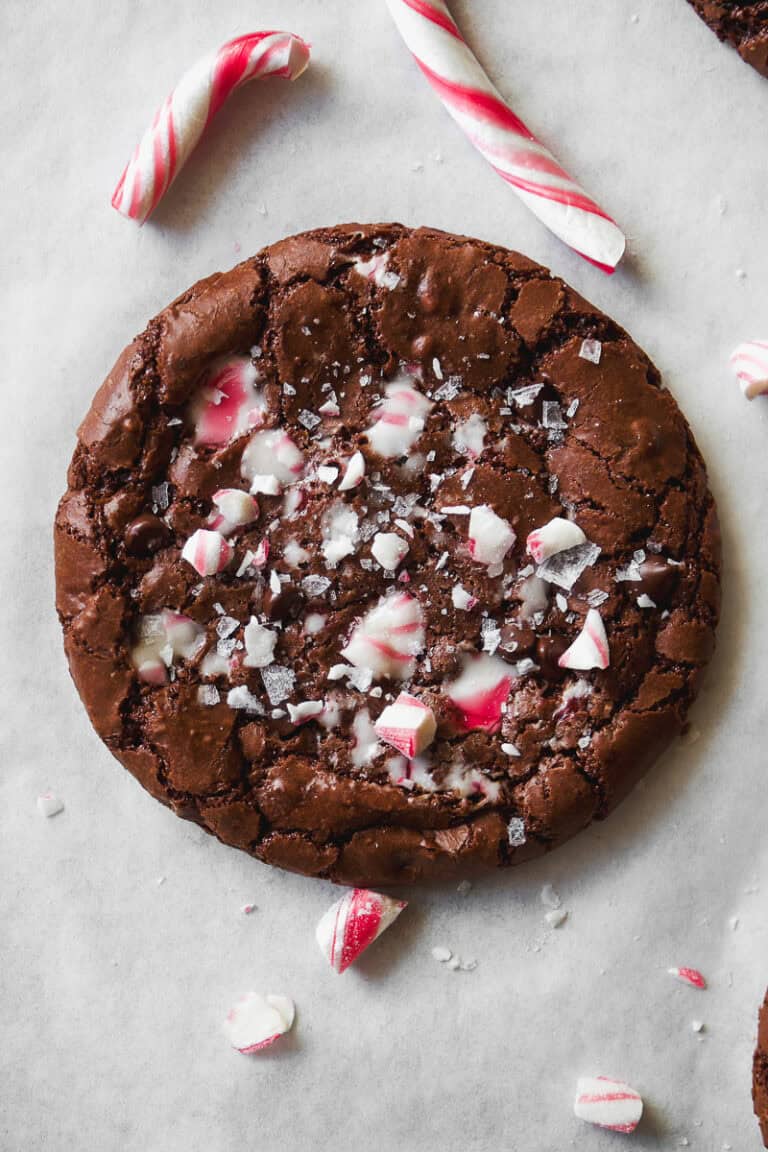 A chocolate cookie topped with crushed peppermint candy pieces sits on parchment paper, surrounded by broken candy canes.