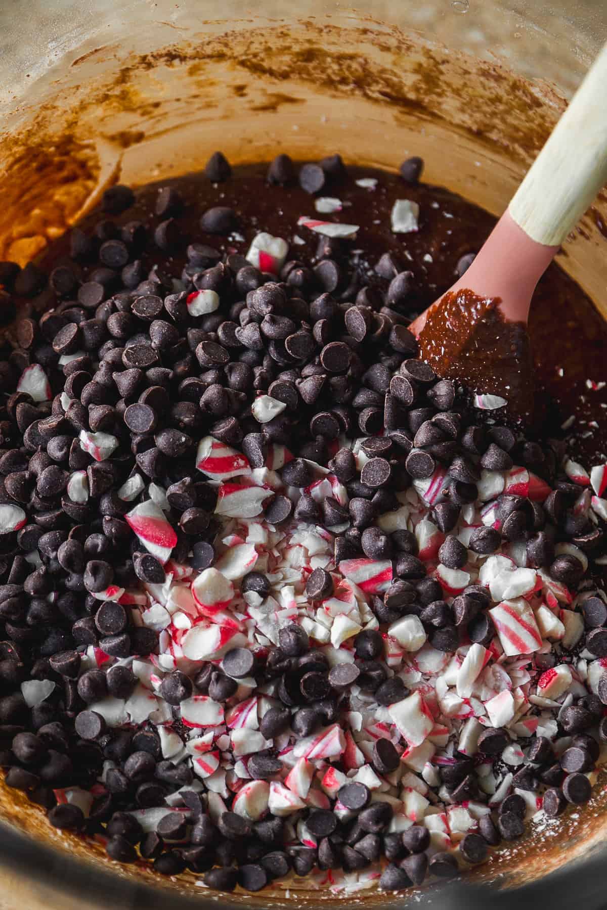 A mixing bowl filled with brownie batter, chocolate chips, and crushed peppermint candies, being stirred with a spatula.