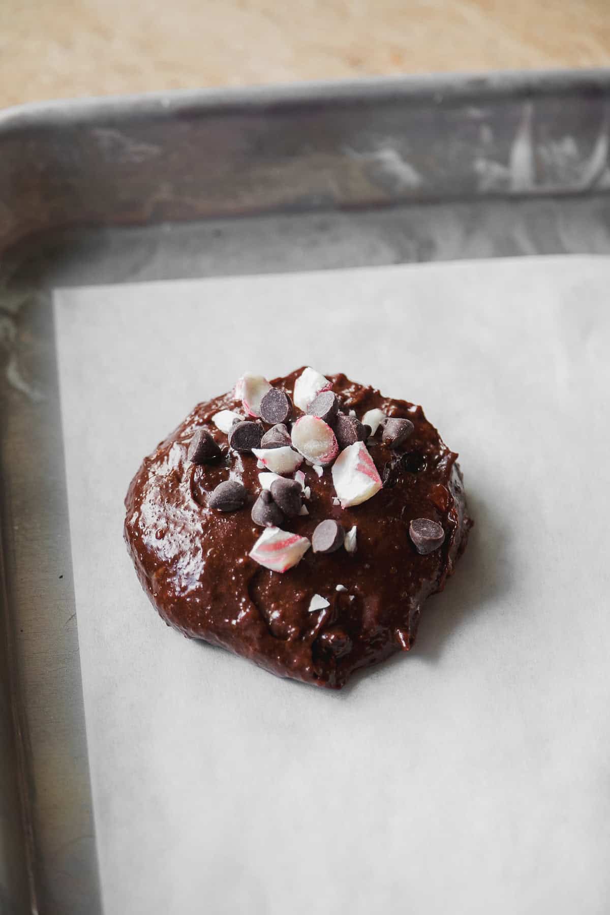 A raw chocolate cookie topped with chocolate chips and crushed peppermint candy sits on a parchment-lined baking sheet, ready to be baked.
