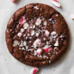 A chocolate cookie topped with crushed peppermint candy pieces, surrounded by broken and whole candy canes on a white surface.