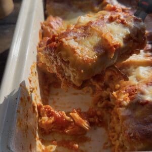 A close-up of cheesy, saucy lasagna roll ups being lifted from a white baking dish, with golden melted cheese and visible layers of pasta and red sauce.