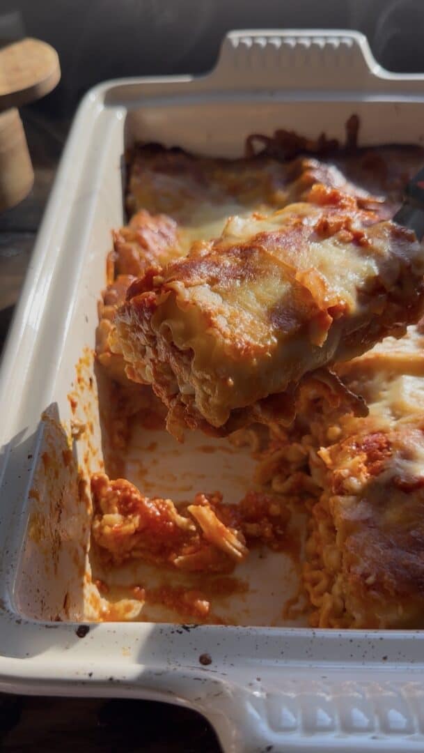 A close-up of cheesy baked lasagna roll ups in a white dish, with one slice being lifted out, showing layers of pasta, sauce, and melted cheese. Sunlight highlights the golden, bubbly top.