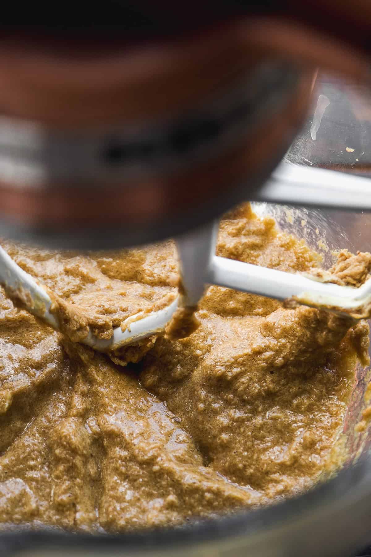Close-up of cookie dough being mixed in a stand mixer with a white paddle attachment, showing a thick, brown batter inside a glass mixing bowl.