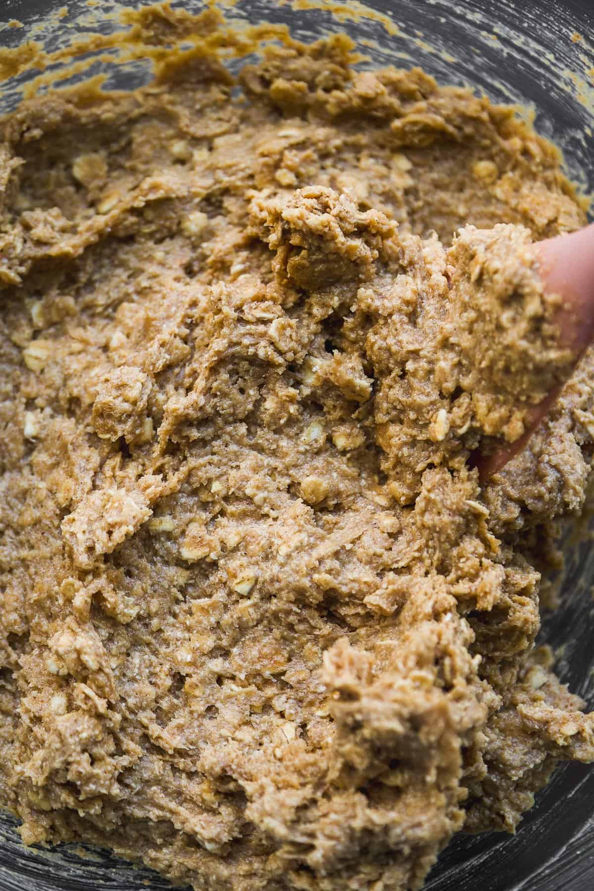 A close-up of thick, textured cookie dough being mixed, showing visible oats and a slightly brown color.