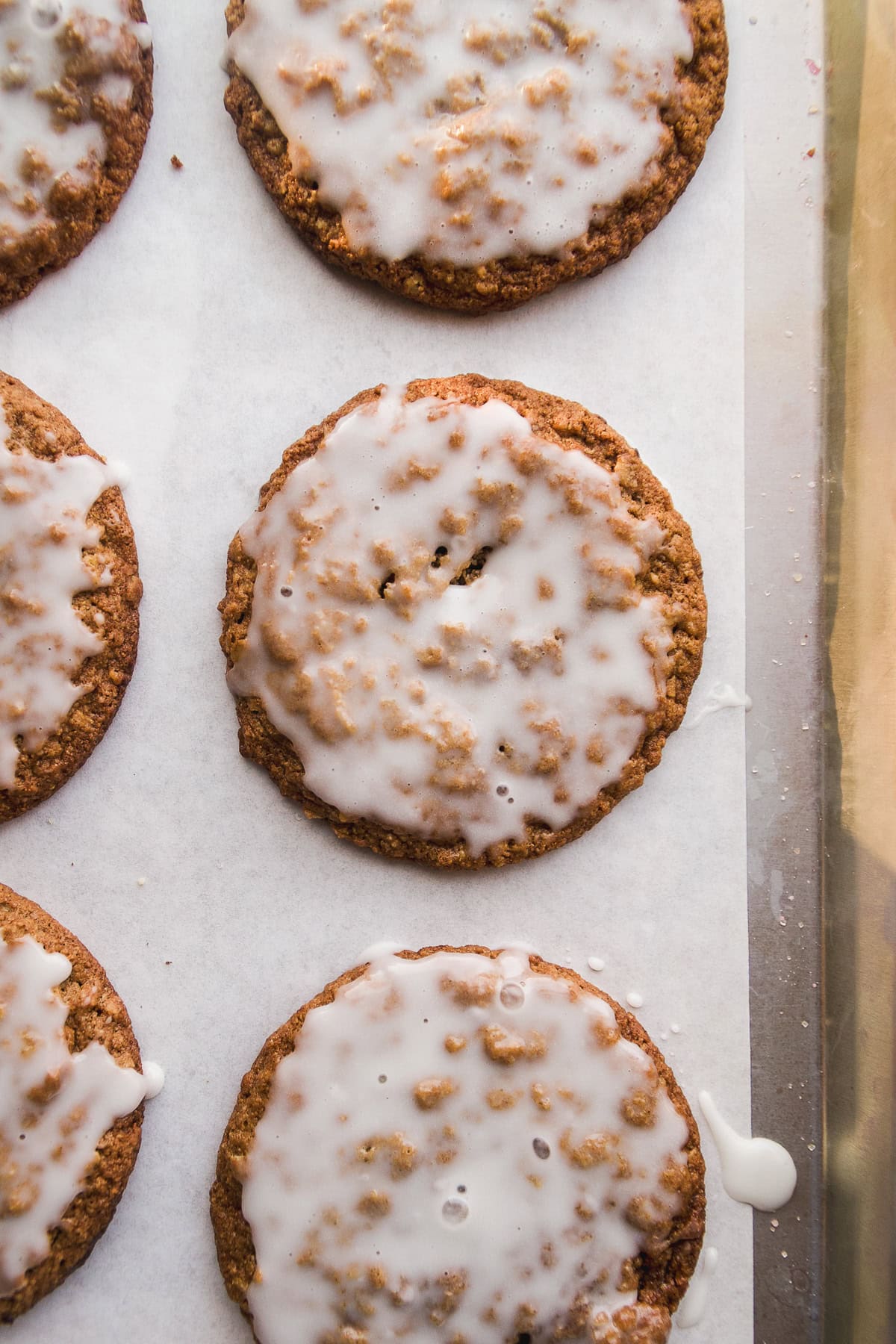 Oatmeal cookies topped with a white sugar glaze are arranged on a sheet of parchment paper, with some glaze dripping off the edges.