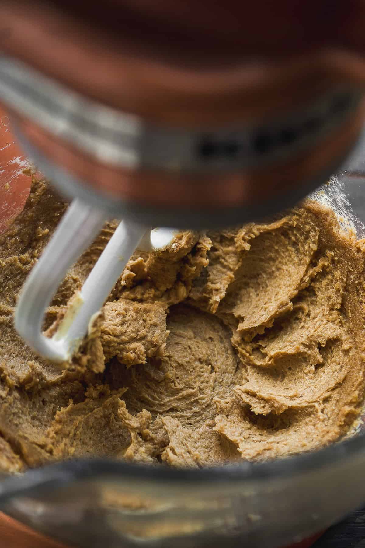 Close-up of a stand mixer with a paddle attachment blending brown cookie dough in a glass mixing bowl.